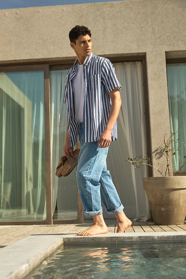 Hombre con jean celeste, camisa rayada de manga corta y remera blanca junto a una pileta