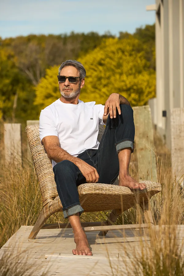 Hombre con remera blanca de manga corta y jean, sentado en un sillón de mimbre